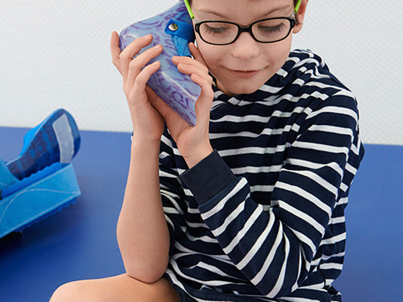 Junge mit Unterschenkelorthese sitzt auf einer Therapieliege und hält sich ein blaues Orthesenteil ans Ohr