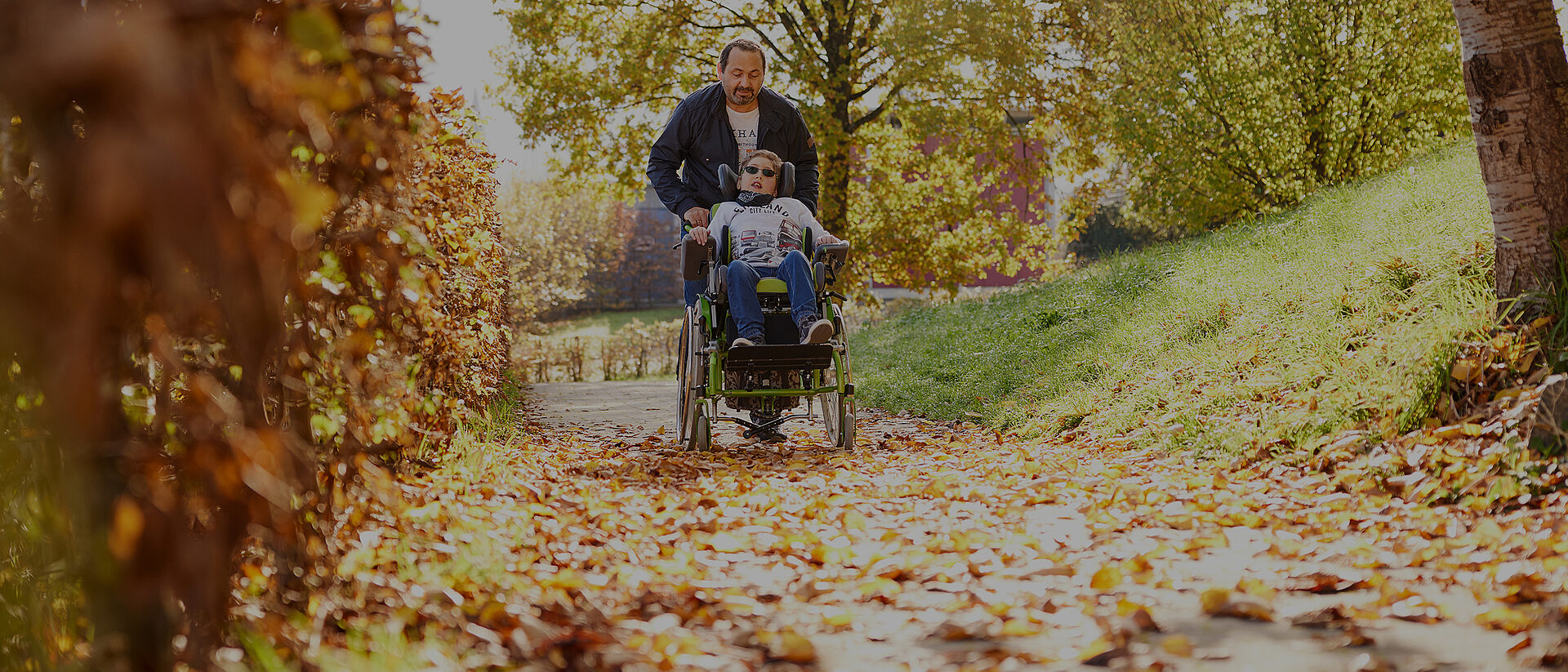 Mann schiebt ein Kind im Reha-Buggy über einen herbstlichen Waldweg