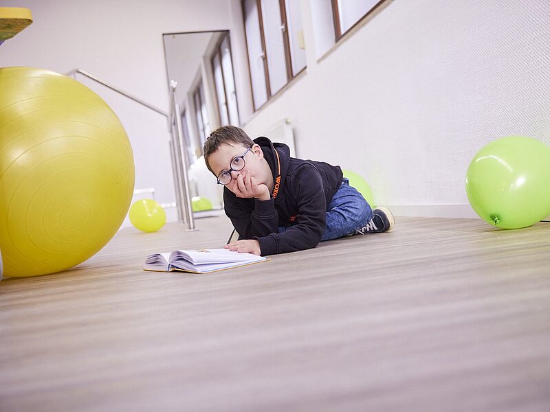 Junge mit Down-Syndrom und Brille liegt lachend neben einem grünen Gymnastikball