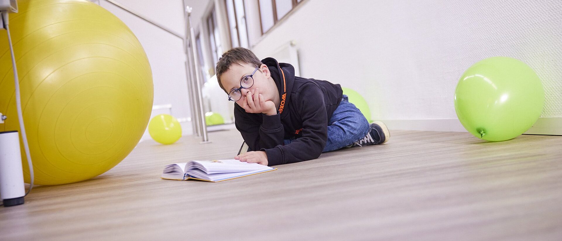 Junge mit Down-Syndrom und Brille liegt lachend neben einem grünen Gymnastikball