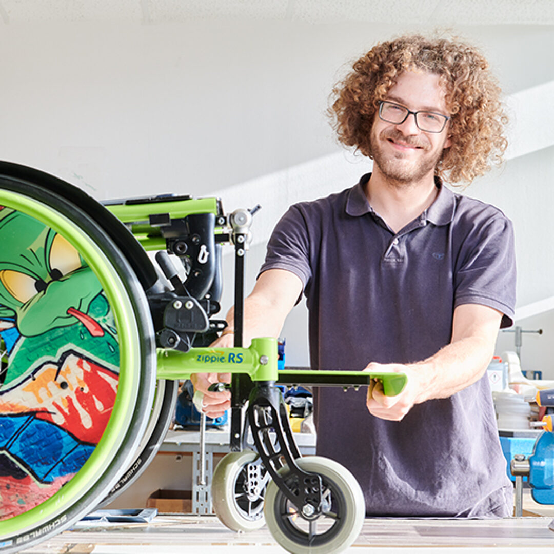 Rehatechniker montiert ein Rad an einen grünen Kinderrollstuhl mit Comic-Speichenschutz