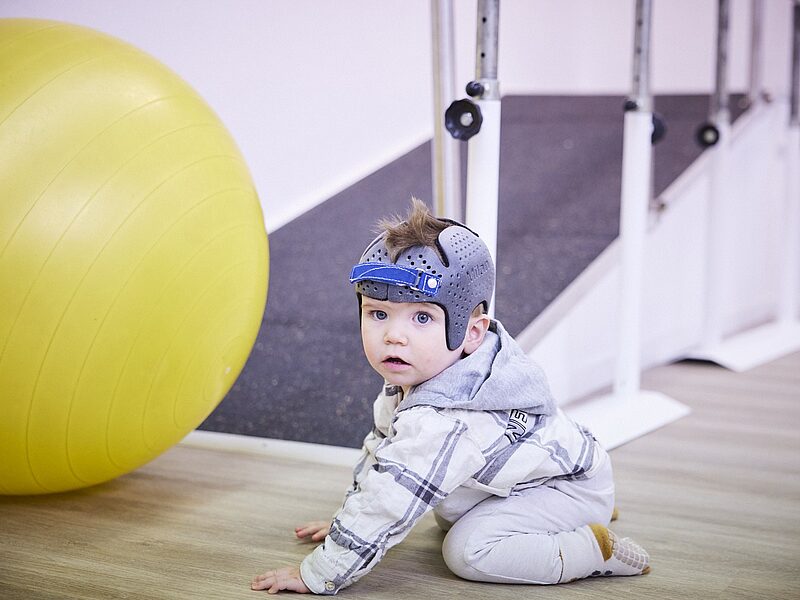 Baby mit Kopforthese sitzt entspannt auf dem Boden im Therapieraum