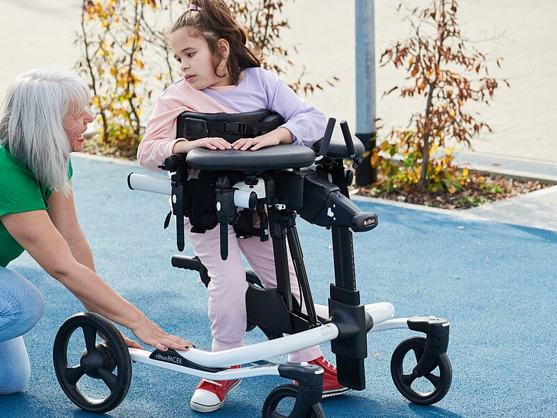 Therapeutin kniet neben einem Mädchen im Gehtrainer auf dem Sportplatz