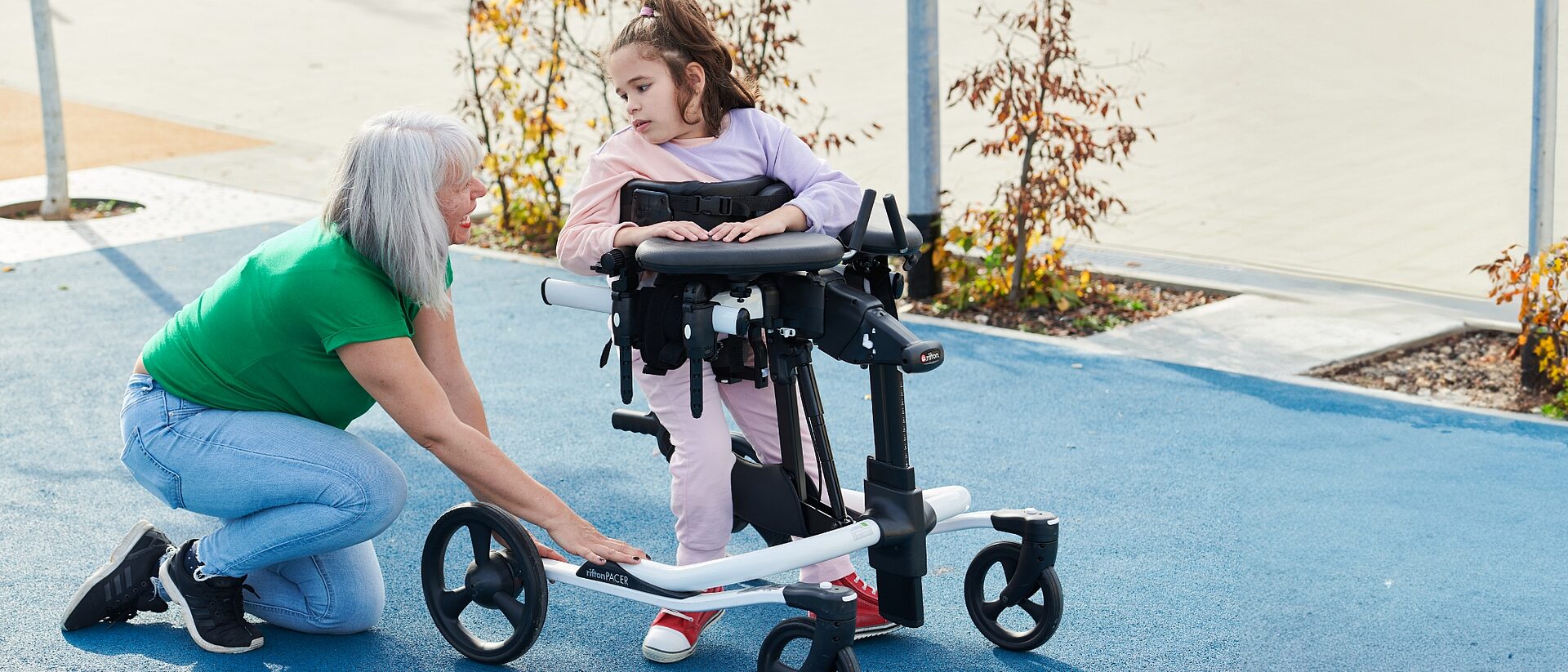 Therapeutin kniet neben einem Mädchen im Gehtrainer auf dem Sportplatz