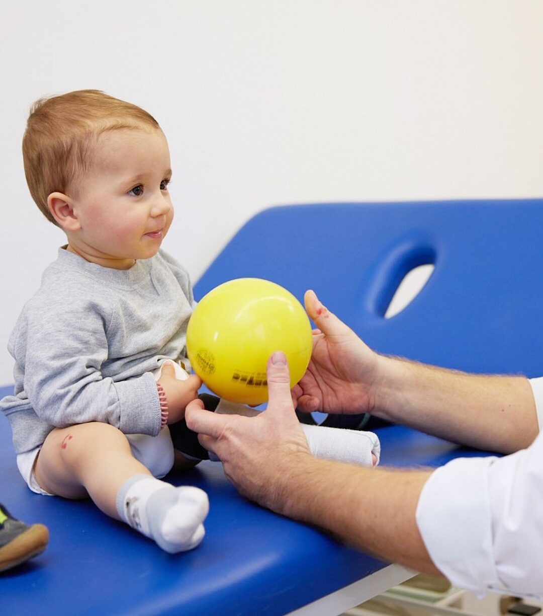 Baby sitzt auf der Therapieliege und hält mit dem Therapeuten einen gelben Ball